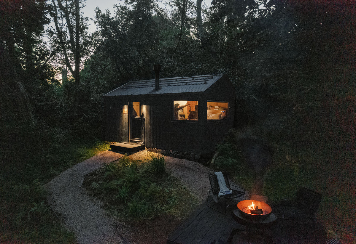 A dark wooden cabin nestled in a dense forest at dusk, with warm lights glowing from the windows and a small fire burning in a fire pit on a deck in the foreground.