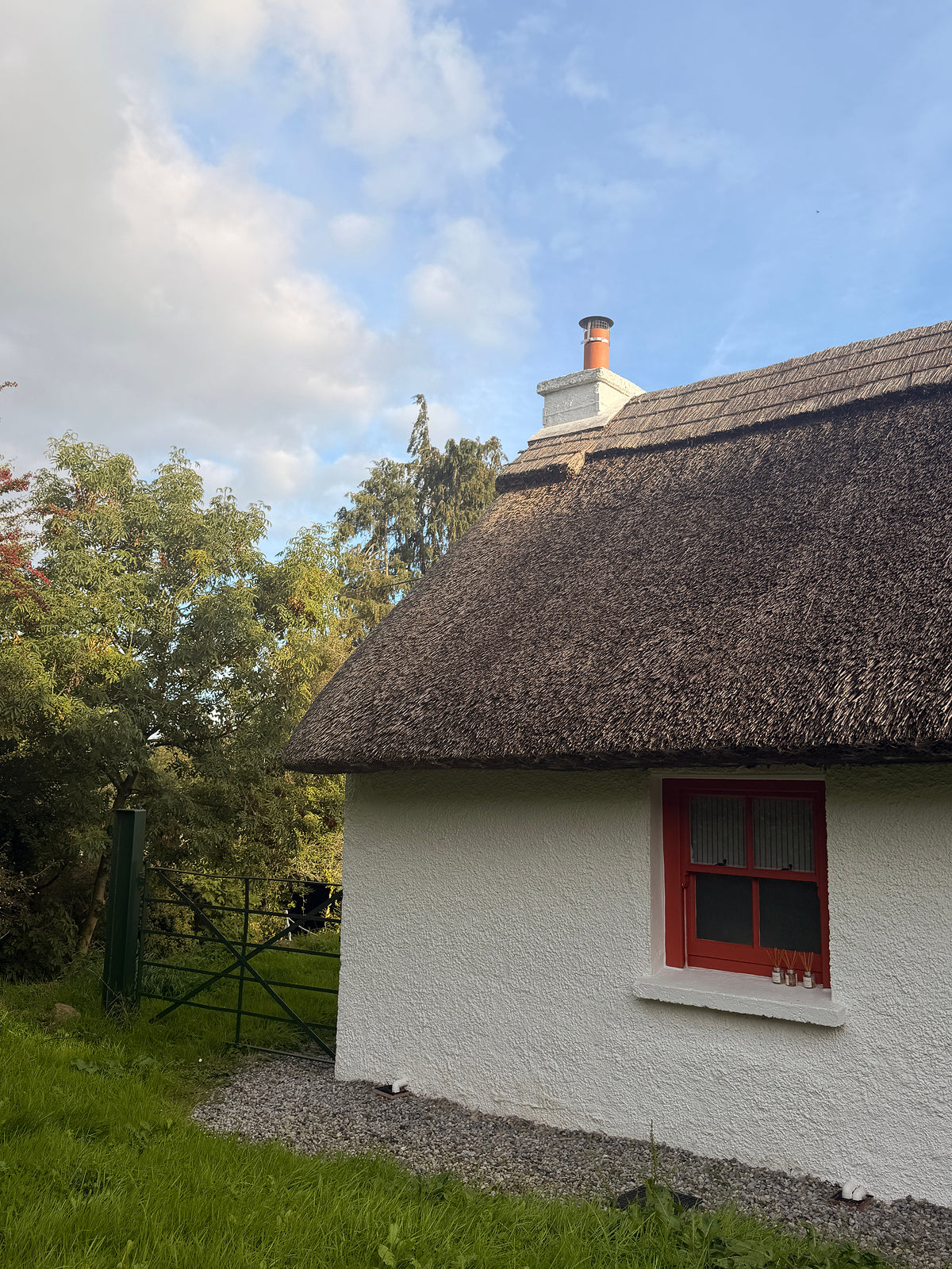 A white cottage with a dark thatched roof and a red-framed window, shown outdoors with grass, trees, a gate, and blue sky in the background.